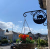 Badger Hanging Basket Bracket A Badger Hanging Basket Bracket by Black Fox Metalcraft Ltd holds a vibrant hanging basket filled with orange flowers on a sunny day, as houses line the street against a clear blue sky.