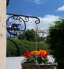 Otter Hanging Basket Bracket An Otter Hanging Basket Bracket by Black Fox Metalcraft Ltd gently swings a basket of red and orange flowers beneath a metal sign featuring a dinosaur silhouette, all illuminated in the sun.