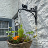 Sheep and Collie Hanging Basket Bracket Beside a small window, the Sheep and Collie Hanging Basket Bracket by Black Fox Metalcraft Ltd is mounted with a hanging flower basket. It's crafted using traditional blacksmithing techniques and features elegant animal silhouettes in a Border Collie design.