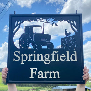 The Tractor Hanging Sign by Black Fox Metalcraft Ltd features "Springfield Farm" with tractor and rooster silhouettes on a fence, displayed outdoors and mounted on a hand-forged bracket for a bespoke touch.