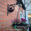 The Badger Hanging Basket Bracket by Black Fox Metalcraft Ltd, skillfully crafted using traditional blacksmithing techniques, supports a hanging basket of flowers on a brick wall. Logo and website visible.