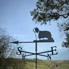 A Bearded Collie Weathervane from Black Fox Metalcraft Ltd, with a rust-resistant finish, stands in a rural landscape under a clear sky, showcasing compass directions.