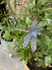 The Metal Dragonfly Ornament by Black Fox Metalcraft Ltd rests among green leaves in a potted outdoor plant, making a charming gift for gardening enthusiasts.