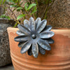 The Flower Plant Pot Hanger by Black Fox Metalcraft Ltd features textured metal petals attached to a terracotta pot, bringing charm as green plants flourish inside.