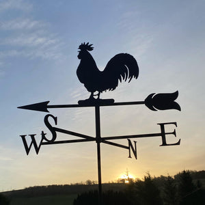 The Cockerel Weathervane from Black Fox Metalcraft's Cockerel weathervane is silhouetted against the morning sunrise, with its traditional directional letters W, S, N, and E clearly seen. In the background, trees and a hill enhance the enchanting scene.