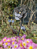 The Hand Crafted Metal Flowers by Black Fox Metalcraft Ltd feature a gilded 23.5-carat, handmade metal sculpture set among pink and yellow primroses, with vines and a wire fence in the background.