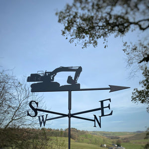 The Doosan Excavator Weathervane by Black Fox Metalcraft Ltd elegantly displays cardinal directions against a rural landscape, with trees framing the scene under a clear blue sky. It features rust protection to ensure lasting durability.