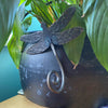 The Dragonfly plant pot hanger by Black Fox Metalcraft Ltd features a hand-forged curled tail design that attaches to your planter—surrounded by lush green leaves, it makes an ideal gift for gardeners.