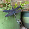 The Dragonfly plant pot hanger by Black Fox Metalcraft Ltd is attached to the rim of a green pot beside ferns and another ceramic pot against a brick wall—an ideal gift for gardeners.