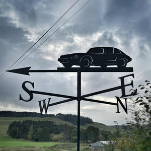 The E Type Jag Weathervane (Series 3) by Black Fox Metalcraft Ltd features a car silhouette atop directional letters S, W, E, and N, with a cloudy sky and rural landscape in the background.