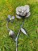 The Hand Crafted Metal Flowers by Black Fox Metalcraft Ltd features two lifelike blooms with layered petals, yellow centers, and 23.5 carat gilded accents, lying on green grass for an elegant touch.
