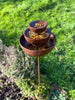 The Black Fox Metalcraft Ltd Insect Bath features three rusted metal bowls stacked on a pole, filled with water and leaves, standing in a grassy outdoor area.