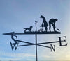 A weathervane from Black Fox Metalcraft Ltd, named "Gardening Couple with Dog," is silhouetted against the sky featuring farm figures: a dog, a bird, a woman with a shovel, and a man gardening. The cardinal directions S, W, N, E are visible.
