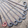 Curved metal rods with spiral ends are displayed on textured stone, reminiscent of bespoke designs. The Whippet Dog Hanging Basket Bracket from Black Fox Metalcraft Ltd is heated and glowing orange at its tip.