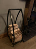 The Log Basket - Blacksmith Made by Black Fox Metalcraft Ltd holds several pieces of firewood beside a fireplace, with fireplace tools on a stand nearby.