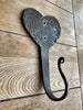 The Hand Forged Heart Hook by Black Fox Metalcraft Ltd is a black metal wall hook with a hammered heart-shaped backplate, shown mounted on wood.