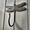 The Hand Forged Dragonfly Hook by Black Fox Metalcraft Ltd is mounted on a vertically paneled wooden surface.