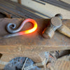 A Fire Poker by Black Fox Metalcraft Ltd, featuring a decorative curl, rests heated on an anvil beside a hammer, showing ongoing metalwork.