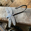 The Hand Forged Dragonfly Hook by Black Fox Metalcraft Ltd, featuring textured wings, rests on an anvil.