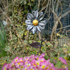 The Forged Metal Flower by Black Fox Metalcraft Ltd features a yellow center, set among pink primroses, ivy, and bare vines—creating a striking steel garden decoration.