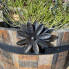 The Flower Plant Pot Hanger by Black Fox Metalcraft Ltd decorates a wooden barrel planter filled with green succulent plants.