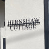 The Laser Cut Metal Sign by Black Fox Metalcraft Ltd, reading "HERNSHAW COTTAGE," is mounted on a white wall, casting a distinctive shadow—an elegant option for house name signs.