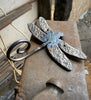 The Metal Dragonfly Ornament by Black Fox Metalcraft Ltd features textured wings and a coiled tail, making it a charming gift for gardeners or nature lovers. Displayed on a workbench, it's perfect for adding artistic flair to any space.