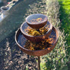 The Black Fox Metalcraft Ltd Insect Bath features three stacked rusted metal bowls of decreasing size on a pole, filled with small stones and set in a garden with soil and grass in the background.