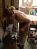 A person wearing an apron is hammering a heated metal rod on an anvil in a workshop with a forge in the background, crafting the Hand Forged Ivy Bracket by Black Fox Metalcraft Ltd.