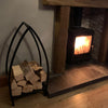 A Black Fox Metalcraft Ltd Log Basket - Blacksmith Made, stacked with firewood, sits on a tile hearth beside a wooden mantel and stone wall.