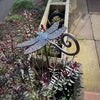 The Metal Dragonfly Ornament by Black Fox Metalcraft Ltd rests among leafy plants in a raised stone planter beside a paved area, making it an ideal gift for any gardener.