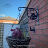A hanging basket with plants is displayed on Black Fox Metalcraft Ltd's Otter Hanging Basket Bracket, which is handcrafted from wrought iron and attached to a brick wall. The background captures part of a building and the sky.