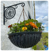 A hanging basket filled with marigolds and various other flowers hangs from the Manchester Bee Bracket by Black Fox Metalcraft Ltd. Mounted against a white wall, it stands out against the cloudy sky and lush green landscape in the background.