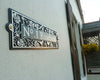 A Metal Art Address Sign from Black Fox Metalcraft Ltd is mounted on a white exterior wall beside a window and a basket of yellow flowers, custom displaying “No. 1 Rosebank.”.