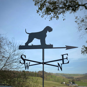 A Schnauzer Weathervane by Black Fox Metalcraft Ltd, featuring a zinc electroplated finish, is silhouetted against a rural landscape with trees and hills in the background.