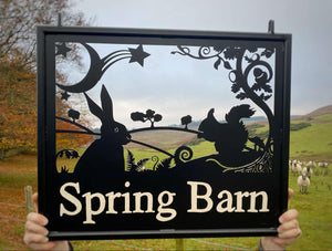 A decorative hanging sign from Black Fox Metalcraft Ltd, featuring silhouettes of a hare and squirrel, reads "Spring Barn" and perfectly blends into the rural landscape with hills and sheep, making it an appealing choice for any nature lover.