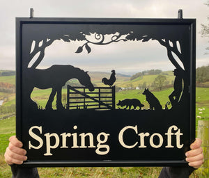 A wrought iron Farm Scene Hanging Sign from Black Fox Metalcraft Ltd, featuring the words "Spring Croft" and decorated with silhouettes of farm animals and a horse, is displayed against the backdrop of rolling hills and trees.