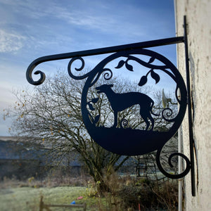 The Whippet Dog Hanging Basket Bracket by Black Fox Metalcraft Ltd features a custom wrought iron silhouette of a hound on a branch, elegantly mounted on a white building. The background includes scenic trees and fields, harmoniously merging art with nature.