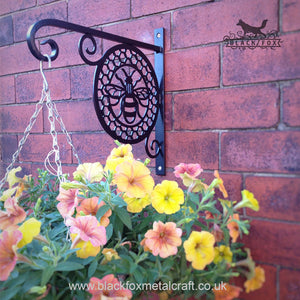 The Manchester Bee Hanging Basket Bracket by Black Fox Metalcraft Ltd adorns the brick wall, holding a basket of yellow and peach flowers. Hand-forged in a traditional coal forge, it beautifully blends artistry with functionality.