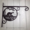 The Otter Hanging Basket Bracket by Black Fox Metalcraft Ltd is a hand-forged ornate metal bracket with a stylized otter design, ideal for displaying your favorite hanging basket and adding charm to any space when mounted on a light gray wooden wall.