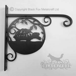 The Tortoise Hanging Basket Bracket from Black Fox Metalcraft Ltd is a black metal wall bracket with a silhouetted tortoise and hare design, featuring decorative scrollwork. This hand-forged garden accessory includes the brand logo, making it perfect for hanging baskets.